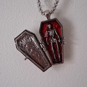 Halsband med skelett i kista - Unikt halsband med ett hängsmycke i form av en kista som kan öppnas. Inuti finns ett detaljerat skelett mot en röd bakgrund. Kedjan och kistan är i silverfärgad metall, vilket ger en cool och mystisk look.