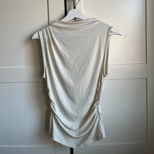 Beige ärmlös topp - Säljer en stilren beige ärmlös topp med draperade detaljer på sidorna. Perfekt för en minimalistisk look. Toppen har en mjuk och följsam passform.