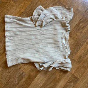 Beige ribbad topp från Gina Tricot - Säljer en stilren beige ribbad topp från Gina Tricot. Toppen har korta ärmar med volangdetaljer som ger en extra touch. Perfekt för en chic och avslappnad look.