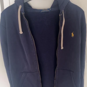 Blå hoodie från Ralph Lauren - Säljer en snygg blå hoodie från Ralph Lauren med dragkedja och huva med snörning. Den har en broderad logga på bröstet och är perfekt för en avslappnad stil. Hoodien är svår att hitta i dagsläge.