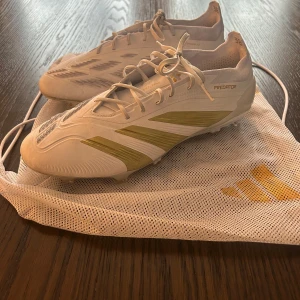 Adidas Predator Elite FG fotbollsskor - Säljer ett par vita Adidas Predator fotbollsskor med guld detaljer. Skorna har snörning och är designade för optimal prestanda på planen. Perfekta för fotbollsspelare som vill ha stil och funktionalitet. Skorna är använda 1 gång och är i utmärkt skick. 