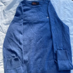 Blå merinoulltröja från Morris - Säljer en stilren blå tröja från Morris i 100% merinoull. Tröjan har en liten broderad detalj på bröstet och långa ärmar med ribbade muddar. Perfekt för våren! Skriv till mig om flera bilder eller frågor💛