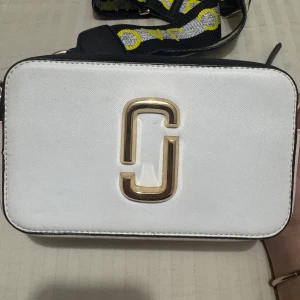 Vit axelväska - Snygg vit axelväska med guldiga detaljer och en bred axelrem med text. Har fått i födelsedagspresent för ca 2 år sen och har använts väl. Dock inga synliga skador/märken