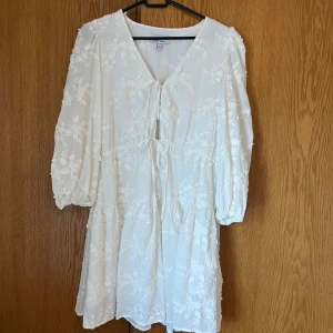 Kort vit klänning  - Superfin vit klänning med blommigt mönster och knytband framtill. Den har trekvartsärmar och  v-ringning. Hade på studenten och älskade. 