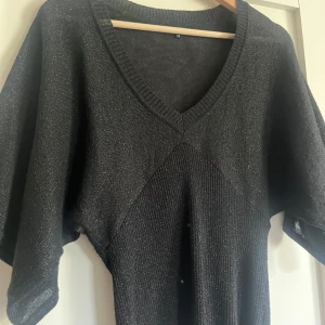 Svart tröja - Snygg svart stickad tröja med v-ringning och korta, vida ärmar. Tröjan är lite glittrig och ribbade detaljer vid halsringningen och nedtill. Perfekt för dig som gillar en chill men ändå stilren look.