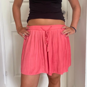 Plisserad korallrosa kortkjol med snörning - Superfin plisserad kortkjol i en härlig korallrosa färg. Kjolen har en luftig känsla, elastisk midja och snygg snörning med tofsar framtill. Perfekt för varma dagar och ger en chill vibe till din outfit. Kjolen har dessutom inbyggda shorts så man känner sig mycket bekväm med den på!! Skriv om du undrar något💕