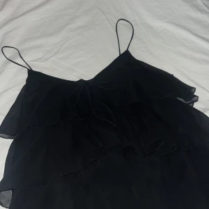 Svart volangig linnetopp med tunna band - Supersöt svart linnetopp med tunna axelband och flera lager volanger. Toppen har en liten knytning framtill och är luftig i modellen, perfekt för varma dagar eller när du vill känna dig extra stylish.