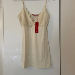 Champagnefärgad linne O.I.S - Söt linne från O.I.S i champagnefärg med tunna axelband och v-ringning. Överdelen har fina spetsdetaljer och broderade blommor, vilket ger en romantisk känsla. Mjuk och lätt i materialet, perfekt för varma dagar.