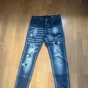 Dsquared2 blå distressed jeans - Säljer ett par blå jeans från Dsquared2 med coola slitningar, färgstänk och tydlig washed effekt. Jeansen har klassisk femficksdesign, låg midja och smal passform. Materialet är bomull och de har en snygg Dsquared2-patch bak.
