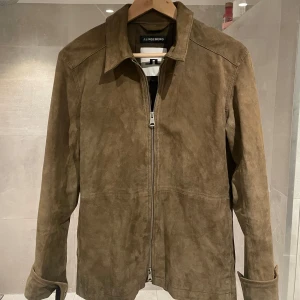 Brun mockajacka från J.Lindeberg - Snygg brun mockajacka från J.Lindeberg med klassisk krage och dragkedja framtill. Jackan har långa ärmar med knäppning vid ärmslut och en stilren, minimalistisk look. Perfekt för dig som gillar en clean och tidlös stil.