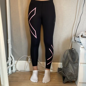 Svarta 2XU tights med rosa detaljer - Säljer ett par svarta tights från 2XU i storlek L. De har snygga rosa grafiska detaljer längs benen och är tillverkade i ett stretchigt syntetmaterial som sitter tight och bekvämt. Perfekta för träning eller löpning.