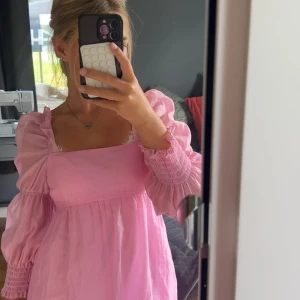 Rosa puffärmklänning med rosett - Supersöt rosa klänning med puffiga långärmar, fyrkantig urringning och stor rosett i ryggen. Klänningen har öppen rygg och är gjord i ett lätt, tunt material som känns luftigt.