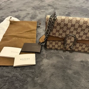 Gucci Dionysus axelväska beige/brun - Säljer en Gucci Dionysus axelväska i beige och brunt GG-monogram. Väskan har silverfärgad kedjerem, ikoniskt spänne framtill och detaljer i mocka. Insidan är fodrad i vinröd sammet. Kommer med dustbag och äkthetskort.
