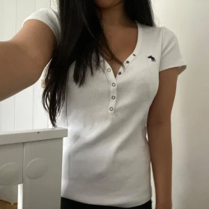 Vit ribbad topp från Abercrombie - Vit ribbad topp från Abercrombie & Fitch med korta ärmar och djup v-ringning med knappar framtill. Liten mörkblå logga broderad på bröstet. Stretchigt bomullsmaterial som sitter snyggt och bekvämt.