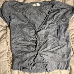 Grå kortärmad knäppblus - Grå kortärmad blus med knappar framtill. Topp i mjukt och tunt material, perfekt för lager-på-lager eller som den är. Enkel och stilren design med ribbade kanter vid ärmslut och nederkant.