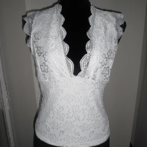 Vit spetsblus med öppen rygg - Supersöt vit blus i spets med blommigt mönster och djup v-ringning. Toppen har en snygg öppen rygg med dekorativ knäppning. Aldrig använd!