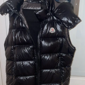 Moncler väst  - Säljer en svart, glansig dunväst från Moncler med klassisk logga på bröstet. Västen har quiltad design, hög krage och dragkedja framtill. Perfekt för lager-på-lager och riktigt snygg till streetwear eller chill outfits.