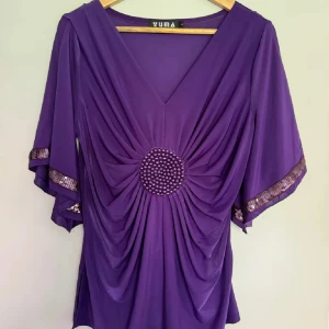 Lila blus med pärlor och paljetter Yuma - Unik lila blus från Yuma med v-ringning, vida ärmar och snygga paljettband längs ärmsluten. Framsidan har ett draperat parti med en rund dekoration av lila pärlor i mitten.