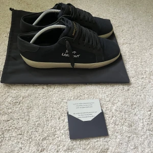 Svarta Saint Laurent sneakers - Snygga svarta sneakers från Saint Laurent med vit text på sidan och gulddetaljer på plösen. Skorna har en klassisk låg profil, svarta snören och en beige platt sula. Ovandelen är i canvas och detaljer i skinn. Perfekta för dig som gillar stilrena och exklusiva sneakers. Box, dustbag och certifikations kort ingår. Skorna är str 44 men passar i 44 1/2 