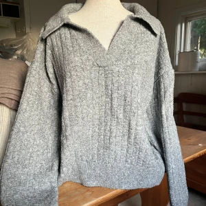 Grå stickad tröja med krage - Mysig grå stickad tröja med bred krage och v-ringning. Tröjan har lång ärm och ribbade muddar nedtill. Perfekt för lager-på-lager och chill dagar. 