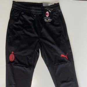 AC Milan träningsbyxor Puma svart S - Svarta träningsbyxor från Puma med AC Milan-logga i rött på ena benet och Puma-logga på andra. Byxorna är i sportig modell med smal passform och tillverkade i polyester. Perfekta för fotbollsträning eller chill. Snygg och modern look.