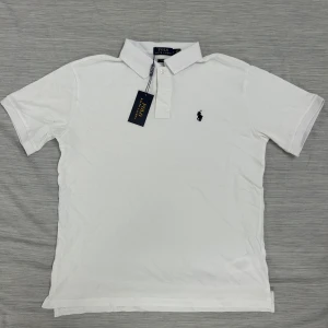 Vit pikétröja från Polo Ralph Lauren - Klassisk vit pikétröja från Polo Ralph Lauren med broderad marinblå logga på bröstet. Tröjan har korta ärmar, krage och knäppning framtill. Tillverkad i mjuk bomull som andas och passar perfekt till en clean och stilren look.