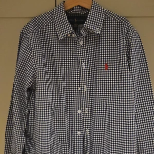 Rutig skjorta från Ralph Lauren M - Snygg rutig skjorta från Ralph Lauren i blått och vitt. Klassisk krage, långa ärmar och knäppning framtill. Ikoniska röda loggan broderad på bröstet. Perfekt för dig som gillar stilrena och tidlösa plagg.