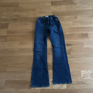 Blå bootcut jeans från Lindex - Snygga blå jeans från Lindex i bootcut-modell med klassisk femficksdesign och knapp i kopparfärg. Jeansen har en bekväm passform och är tillverkade i slitstarkt denimtyg. Perfekta för dig som gillar en retrovibe och vill ha ett par jeans som sticker ut.