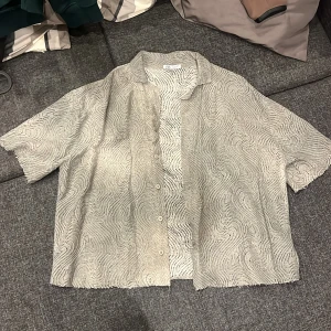 Grå mönstrad skjorta från Zara - Säljer en grå skjorta från Zara i storlek L med korta ärmar och coolt vågigt mönster. Skjortan har knappar framtill och råa kanter vid ärmslut och nederkant. Materialet är lätt och luftigt, perfekt för varmare dagar. Snygg och unik design som sticker ut.