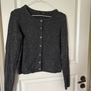 Mörkgrå kofta från 157 med knappar - Mysig mörkgrå kofta från 157 i stickad kvalitet. Koftan har rund halsringning, långa ärmar och lila knappar framtill. Perfekt att slänga på sig när det är kyligt ute. Enkel och stilren design som funkar till det mesta.