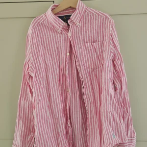 Rosa och vit randig skjorta Ralph Lauren - Snygg rosa och vit randig skjorta från Ralph Lauren i luftig linnemix. Klassisk button-down krage, bröstficka och broderad logga i turkos på ärmen. Perfekt för en fräsch och avslappnad stil.