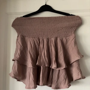 Beige volangkjol med smock - Supersöt kort volangkjol i beige med bred smock i midjan. Kjolen har flera lager volanger som ger en flowy och luftig känsla. Perfekt för dig som gillar en feminin och avslappnad stil.