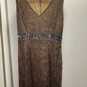 Brun glittrig långklänning med paljetter - Snygg brun långklänning med v-ringning och glittriga detaljer över hela tyget. Midjan pryds av ett band med paljetter och broderade blommor i lila och silver. Perfekt för dig som vill sticka ut med en elegant och unik look.