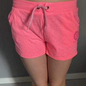 Rosa shorts med snörning från Esprit - Snygga rosa shorts från Esprit med dragsko i midjan och två fickor. Mjukt bomullsmaterial och tryckt logga på ena benet. Perfekta för varma dagar eller chill hemma. Sportig vibe och riktigt skön passform.