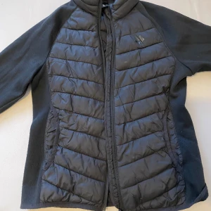 Svart quiltad jacka från Lager 157 - Svart jacka från Lager 157 med quiltade paneler fram och bak samt mjuka ärmar. Jackan har dragkedja hela vägen och en sportig look, perfekt för kyliga dagar. Materialmix av syntet och polyester ger både värme och rörelsefrihet.