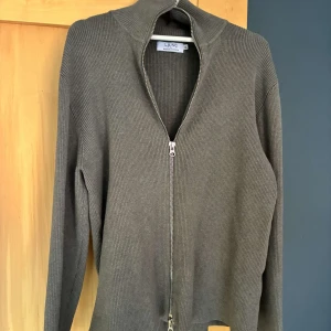 Olivgrön ribbad kofta från Ljung - Säljer en olivgrön ribbad kofta från Ljung med hel dragkedja framtill och hög krage. Tröjan är långärmad och har en clean, minimalistisk look som passar perfekt till lager-på-lager-stil. Materialet är mjukt och stickat för extra komfort.