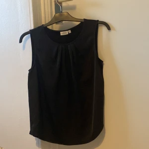 Svart ärmlös blus från KappAhl - Stilren svart blus från KappAhl med rund halsringning och fina veck framtill. Modellen är ärmlös och har en rak passform som passar perfekt till både jeans och kjol. Enkel och snygg design som funkar till många olika looks.