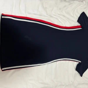 Marinblå kortklänning med ränder - Snygg marinblå kortklänning med sportiga röda och vita ränder längs sidorna. Klänningen har korta ärmar och en figurnära passform som ger en cool och modern look. Perfekt för dig som gillar stilrena och trendiga plagg.