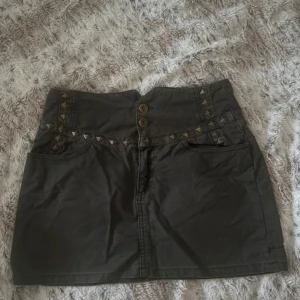 Grön kortkjol med nitar  - Cool grön kortkjol med dubbla knappar framtill. Kjolen har dekorativa nitar i silver längs midjan och fickor både fram och bak. Perfekt för dig som gillar edgy detaljer och vill sticka ut lite extra. Storlek M men passar även mig som är S