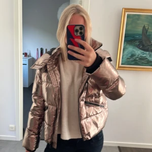 Metallic rosa pufferjacka med huva - Säljer en metallic rosa pufferjacka med huva och dragkedja. Jackan har quiltad design, två snedställda fickor med dragkedja och en cool glansig finish. Perfekt för dig som vill sticka ut och hålla dig varm under kalla dagar.