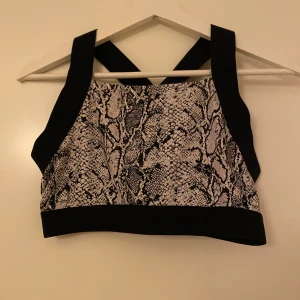 Sporttopp - Sporttopp från Ellos med svart-vitt ormmönster och breda svarta axelband. Toppen har en sportig vibe med croppad passform och är perfekt för att sticka ut. Materialet är syntet och känns lätt och smidigt mot huden. 