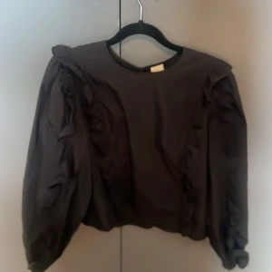 Svart blus med volang från H&M - Säljer en svart blus från H&M i storlek 38. Blusen har långa puffärmar och snygga volangdetaljer längs axlar och ärmar. Den har rund halsringning och en liten öppning bak. Perfekt för dig som gillar trendiga och feminina detaljer.