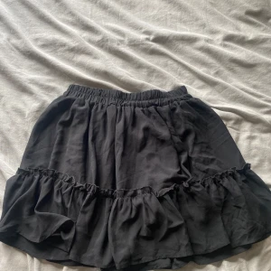 Svart volangkjol med resårmidja - Supersöt svart kortkjol med volang och resår i midjan. Kjolen har en luftig och flowy känsla, perfekt för att styla med en crop top eller oversized hoodie. Enkel men ändå trendig design som passar till sneakers eller boots.