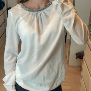 Supersöt vit blus med pärlkrage från Hallhuber💖 - Jättesnygg vit blus från Hallhuber med lång ärm och rund halsringning dekorerad med pärlor och glittriga detaljer. Blusen har en lätt glansig finish och knäppning bak i nacken. Perfekt för dig som vill ha något stilrent men ändå extra.