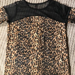 Leopardmönstrad mesh-topp - Cool topp med leopardmönster i beige och svart. Överdelen och axlarna är i transparent svart mesh som ger en edgy vibe. Korta ärmar och rund halsringning. Perfekt för dig som vill sticka ut med ett djurmönster och trendiga meshdetaljer.