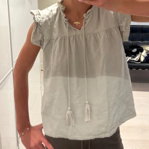 Ljusgrå beige blus med knyt och volang - Supersöt ljusgrå blus med volangdetaljer vid ärmslut och hals, samt långa knytband med tofsar framtill. Blusen är ärmlös med lätt rynk vid axlarna och har en avslappnad passform. Perfekt för en chill och stilren look. Från only passar mig som är 169,5