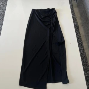 Svart långkjol med slits och rynk - Svart långkjol i mjukt material med snygg rynkdetalj i midjan och en stilren slits framtill. Kjolen har en omlottkänsla och passar perfekt till sneakers eller boots för en cool look.