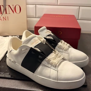 Valentino Garavani Open sneakers vit/svart - Snygga Valentino Garavani Open sneakers i vitt skinn med bred svart läderdetalj över vristen och ikoniska nitar baktill. Klassisk rund tå, vita snören och chunky sula. Kommer med originalkartong och dustbag. Perfekta för dig som gillar lyxig streetstyle.