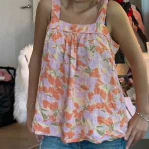 Blommig linnetopp i pastellfärger - Supersöt linnetopp med breda axelband och blommigt mönster i orange, lila, grönt och lite gult. Luftig och flowy passform, perfekt för varma dagar. Toppens material känns lätt och svalkande, och färgerna ger en riktigt somrig vibe.