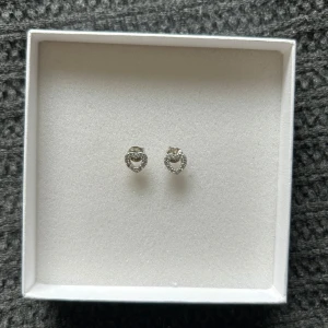 Hjärtformade örhängen i silver - Supersöta små örhängen i form av hjärtan, tillverkade i silverfärgat material och dekorerade med glittrande stenar runt hela hjärtat. 